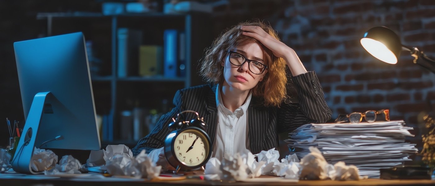 A stressed worker at a cluttered desk at night beside an alarm clock and crumpled paper